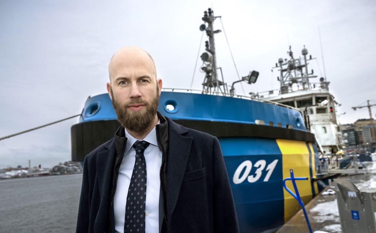 Minister för civilt försvar Carl Oskar Bohlin (M) i halvbild framför ett fartyg från kustbevakningen.